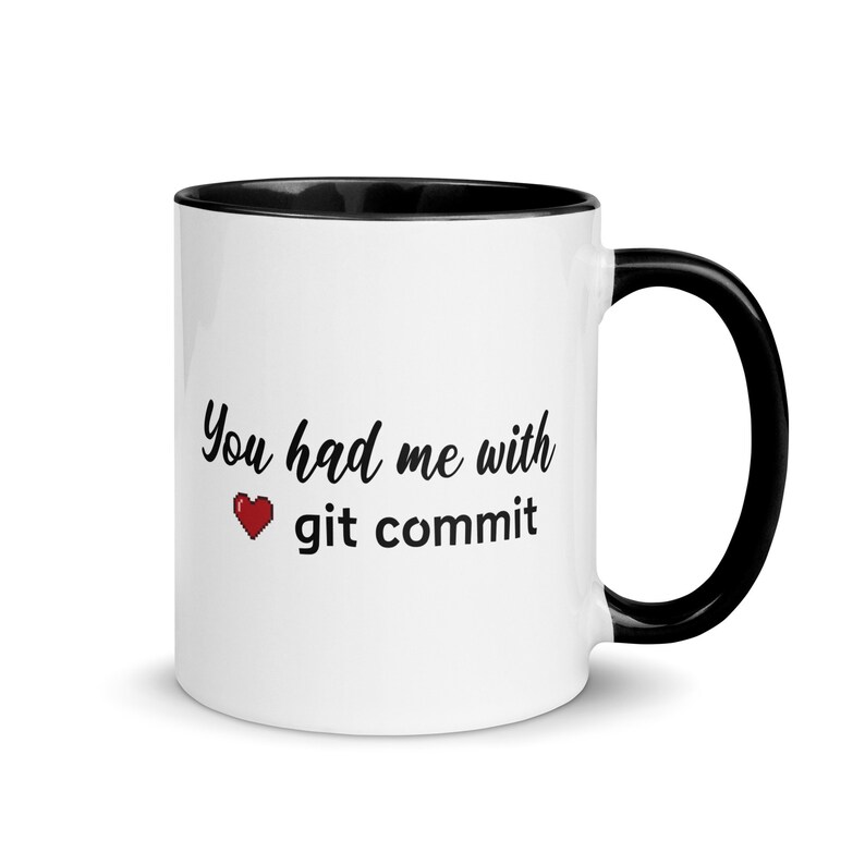Op de afbeelding: Een witte koffiemok met een zwarte rand en handvat. De mok heeft een zwart-witte afbeelding met de tekst "You had me with &hearts; git commit".