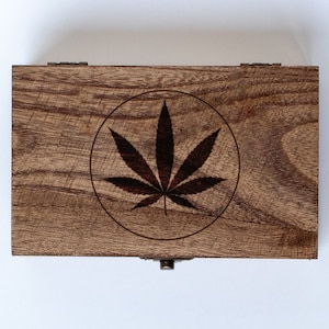 May include: A wooden box with a hinged lid and a brass latch. The box has a circular design with a black marijuana leaf inside.