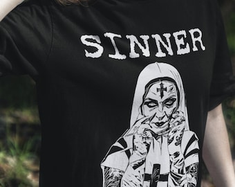 May include: A woman wearing a black t-shirt with the word "SINNER" printed on it. The shirt features a graphic of a nun with a cross on her forehead and a cross on her chest. The nun is holding her hands up to her face.