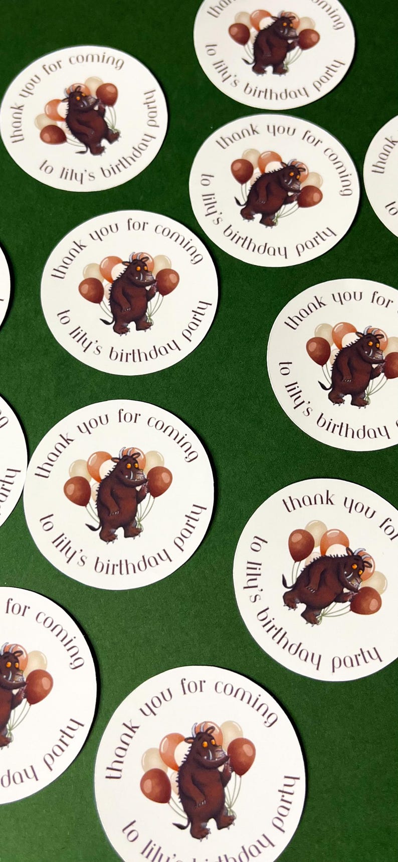 May include: White circular stickers with a brown cartoon creature holding balloons. The stickers say "Thank you for coming to Lily's birthday party" in a circular design. The background is a solid green.