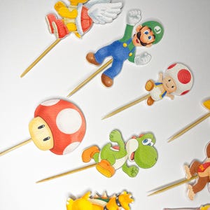 May include: A selection of colourful cupcake toppers featuring cartoon characters on wooden sticks. The toppers depict characters such as Mario, Luigi, Yoshi, and Bowser, with vibrant colours and detailed designs. Ideal for themed parties or celebrations.
