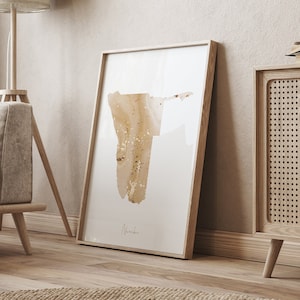 Peut inclure: Une impression encadrée d'un contour doré de la Namibie sur fond blanc. Le cadre est en bois brun clair et l'impression est accrochée à un mur beige. L'impression est dans un salon avec une table d'appoint en bois et une lampe.
