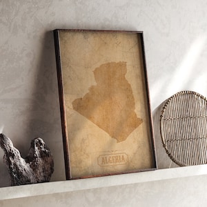 May include: A framed vintage map of Algeria with a brown background and a brown wooden frame. The map is on a white shelf with a woven basket and brushes.