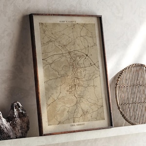May include: Framed vintage map print of Gera, Germany, with coordinates at the top. The map is in sepia tones, showing roads and city layout. The frame is dark brown, and the print is displayed on a white shelf.