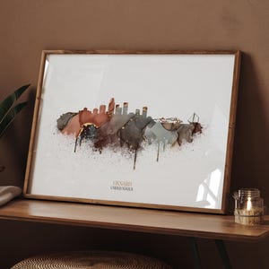 Może przedstawiać: Oprawiony wydruk artystyczny przedstawiający akwarelową panoramę Oxnard, Stany Zjednoczone. Grafika wykorzystuje odcienie brązu, szarości i złota z efektem rozprysku. Wydruk jest prezentowany w drewnianej ramie.