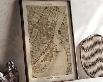 May include: A framed sepia map of Montreal, Canada, showing the city's layout, streets, and waterways. The dark brown frame surrounds the map, which includes coordinates at the top. The map is displayed on a white shelf with a wooden sculpture and a woven circular object.