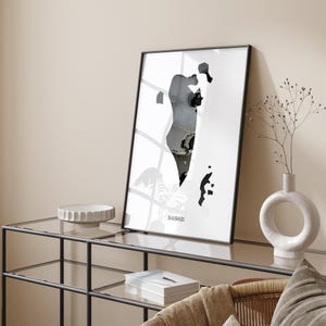 May include: A black and white print of the outline of Bahrain, with the word "Bahrain" printed below the outline. The print is framed in a black frame and is hanging on a wall above a glass-top table.