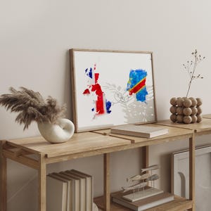 May include: A framed watercolour print depicting the maps of the UK and the Democratic Republic of Congo, linked by a fine line. The artwork is displayed on a wooden shelf with decorative items, including a vase with dried plants and a wooden sphere sculpture.