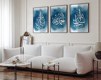 May include: Three framed prints with blue watercolor backgrounds and white Arabic calligraphy. The prints are hung on a white wall above a white couch.