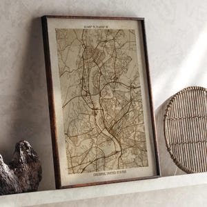 Puede incluir: Una impresión enmarcada del mapa de Chicopee, Estados Unidos, en tonos sepia. El mapa detalla el trazado de la ciudad con calles y vías fluviales. El marco es de color marrón oscuro. Las coordenadas están impresas en la parte superior y el nombre de la ciudad en la inferior.