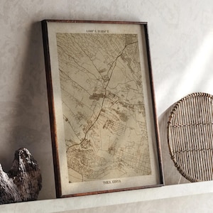 May include: A framed map of Thika, Kenya, in a brown wooden frame. The map is a detailed, sepia-toned illustration of the city's layout, including roads and buildings. The frame is rectangular and the map is displayed on a white surface.