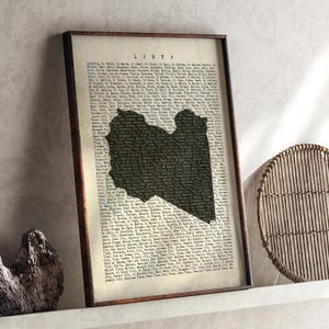Könnte beinhalten: Gerahmter Druck mit einer schwarzen Karte von Libyen, überlagert auf einem Hintergrund aus Text. Das Wort "LIBYEN" steht oben. Der Rahmen ist dunkelbraun. Der Druck wird auf einem weißen Regal mit einem dekorativen Holzstück und einem gewebten kreisförmigen Objekt ausgestellt.