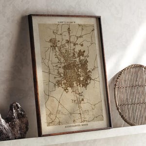 May include: Framed map of Aguascalientes, Mexico, in a dark brown wooden frame. The map is a detailed, sepia-toned illustration of the city's layout, with streets and buildings. Coordinates are printed at the top. The city name is printed at the bottom.