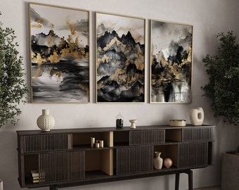 May include: Three framed abstract art prints. Each features a gold frame and a watercolor-style design in black, gray, and gold. The artwork depicts mountain-like shapes with gold accents. The prints are displayed above a dark wood cabinet with decorative items.