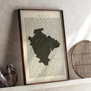 May include: Framed print of the county of Devon, England, with a dark silhouette of the county's shape. The print is set against a vintage-style background with the word "DEVON" at the top. The frame is a dark brown color.