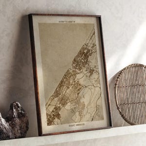 Può includere: Mappa incorniciata di Rabat, Marocco, in tonalità seppia. La mappa mostra strade e caratteristiche geografiche, incorniciata in legno marrone scuro. Le coordinate sono stampate in alto e il nome della città in basso.