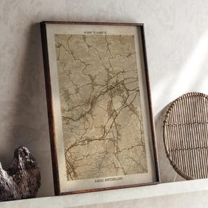 May include: Framed vintage-style map print of Aarau, Switzerland, with coordinates 47.3904° N, 8.0457° E. The map features detailed brown lines on a beige background, encased in a dark brown wooden frame. The print is displayed on a white shelf.