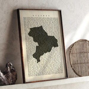 May include: Framed print featuring a black silhouette of Hackney, London, against a backdrop of text. The print is in a dark wooden frame and rests on a white shelf. The word "HACKNEY" is printed at the top.