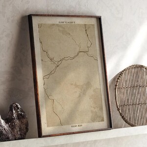 May include: A framed map print of Najaf, Iran, in a brown wooden frame. The map is in sepia tones with the text "32.094° N, 44.321° E" at the top and "Najaf, Iran" at the bottom. The print is displayed on a white shelf.