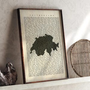May include: A framed print featuring a map of Switzerland in a dark silhouette against a vintage-style background of text. The print is encased in a dark brown wooden frame, adding a classic touch to the artwork. The word "SWITZERLAND" is visible at the top.