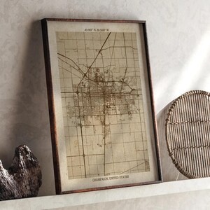 May include: A framed map print of Champaign, United States, in sepia tones. The map shows the city's layout with streets and buildings. The frame is dark brown. The text "40.1603° N, 88.3435° W" and "CHAMPAIGN, UNITED STATES" are visible.