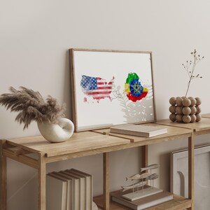 Może przedstawiać: Oprawiony wydruk przedstawiający akwarelowe mapy Stanów Zjednoczonych i Etiopii, połączone cienką linią. Mapa USA jest pokolorowana flagą amerykańską, a mapa Etiopii prezentuje swoje barwy narodowe. Wydruk jest eksponowany na drewnianej półce.