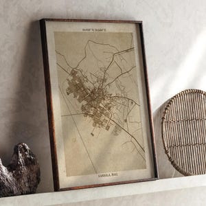 May include: Framed map print of Karbala, Iraq, in sepia tones. The map shows the city's layout with roads and buildings. The frame is dark brown. The text "25.7838° N, 58.8464° E" is at the top, and "KARBALA, IRAQ" is at the bottom.