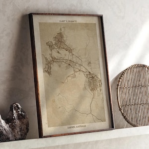 May include: A framed map of Darwin, Australia, in a dark wooden frame. The map has detailed street layouts and geographical features, with coordinates at the top and the city name at the bottom. The color palette is neutral, with a sepia tone, giving it a vintage appearance.