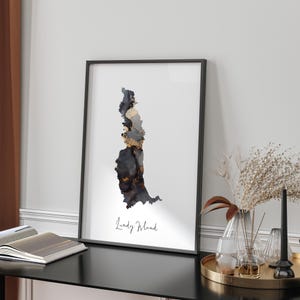 May include: A black and white abstract art print of an island with gold accents. The print is framed in a black frame and the text "Lundy Island" is written below the image.