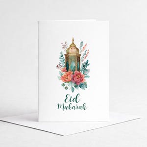 Op de afbeelding: Witte wenskaart met een aquarel illustratie van een gouden lantaarn omringd door roze en oranje bloemen en groen blad. De woorden "Eid Mubarak" zijn in groene letters geschreven.