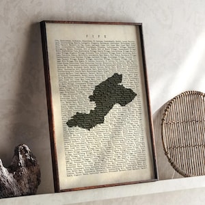 Op de afbeelding: Een ingelijste print met een kaart van Fife, Schotland, in een donker silhouet tegen een vintage-achtige achtergrond van tekst. De print zit in een donkerbruine lijst, met het woord "FIFE" bovenaan.