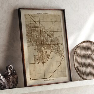 May include: A framed map print of Regina, Canada, in sepia tones. The map shows detailed road networks and city layouts. The frame is a dark brown colour, and the print includes the coordinates 50.4452° N, 104.6189° W.