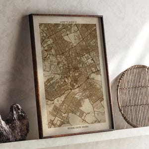 Op de afbeelding: Een ingelijste kaart van Riyad, Saoedi-Arabië, in sepia tinten. De gedetailleerde kaart toont straten en gebouwen. De donkerbruine lijst omringt de kaart. Coördinaten staan bovenaan afgedrukt. De kaart leunt tegen een muur.