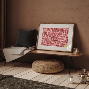 Op de afbeelding: Een ingelijst afdruk van islamitische kalligrafie in rood en goud op een witte achtergrond. De afdruk hangt aan een houten wandplank met een geweven kussen en kaarsen op de vloer.