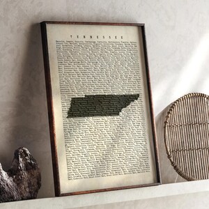 May include: Framed vintage-style print featuring the state of Tennessee. The map of Tennessee is overlaid on a background of text. The frame is a dark brown color.