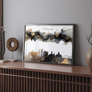 Könnte beinhalten: Gerahmter Kunstdruck mit einer schwarzen Silhouette der Skyline von Middlesbrough, England. Das Kunstwerk enthält goldene und schwarze abstrakte Tintenmuster über der Skyline, mit dem Wort "Middlesbrough" in Schreibschrift. Der Rahmen ist dunkelgrau.