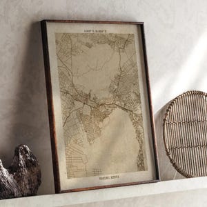 May include: A framed map print of Nakuru, Kenya, in sepia tones. The map details the city's layout with roads and buildings. The frame is dark brown, and the print is displayed on a white shelf. The text "NAKURU, KENYA" is visible at the bottom.