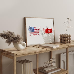 May include: A framed print featuring watercolor maps of the United States and Turkey, with their respective flags incorporated into the designs. The artwork is displayed on a wooden shelf, alongside decorative items like a vase and a wooden sculpture.