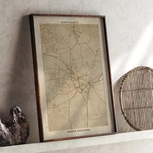 May include: A framed map print of Rangpur, Bangladesh, with coordinates 25.7404° N, 89.2618° E. The sepia-toned map shows a network of roads and streets, set within a dark brown wooden frame. The print is displayed on a white shelf.
