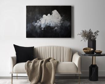 May include: An abstract painting of a cloud in shades of white and gray hangs above a cream-colored sofa with a black pillow and a tan throw. A small side table holds gold bowls and a black vase.