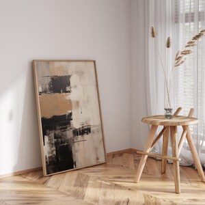 May include: Abstract art painting in shades of brown, black, and white in a wooden frame leaning against a white wall. A wooden stool with a round top sits in the corner of the room.