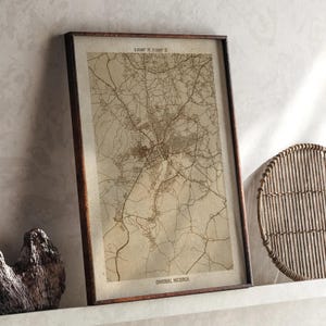 May include: Framed map print of Owerri, Nigeria, in sepia tones. The map features intricate road and city details, with coordinates at the top. The print is encased in a dark brown wooden frame, leaning against a neutral-colored wall.