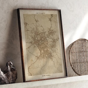 Puede incluir: Una impresión enmarcada de un mapa de Akure, Nigeria, en un marco de madera marrón. El mapa es una representación detallada, en tonos sepia, del diseño de la ciudad, incluyendo carreteras y áreas urbanas. El texto "AKURE, NIGERIA" está en la parte inferior.