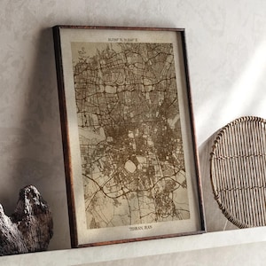 May include: Framed sepia-toned map print of Tehran, Iran. The map features intricate street details and is encased in a dark wooden frame. The city's name is printed at the bottom, with coordinates at the top. The print is leaning against a wall.