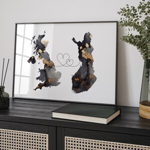 Może przedstawiać: Oprawiony wydruk artystyczny przedstawiający abstrakcyjne mapy akwarelowe Wielkiej Brytanii i Finlandii, połączone linią w kształcie serca. Mapy są w odcieniach czerni, szarości i złota na białym tle. Grafika jest prezentowana na ciemnej drewnianej powierzchni.