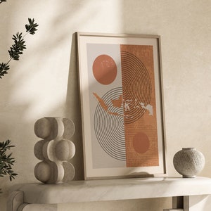 May include: A framed abstract print with a beige background, two orange circles, and black lines forming a circle pattern. The print is on a white marble shelf with a grey stone vase and a grey stone bowl.