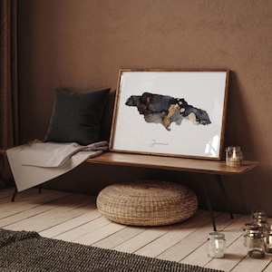 May include: A framed print of a black and gold watercolor map of Jamaica. The map is on a white background with the word "Jamaica" written below it. The print is sitting on a wooden bench with a woven ottoman in front of it.
