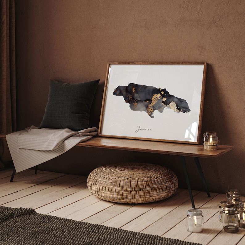 May include: A framed print of a black and gold watercolor map of Jamaica. The map is on a white background with the word "Jamaica" written below it. The print is sitting on a wooden bench with a woven ottoman in front of it.