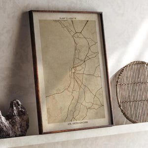 Op de afbeelding: Ingelijste kaartprint van Ayr, Verenigd Koninkrijk, met coördinaten 55.4596° N, 4.6292° W. De kaart toont een netwerk van bruine lijnen op een beige achtergrond, omlijst door een donkerbruine houten lijst. De tekst "AYR, UNITED KINGDOM" staat onderaan.