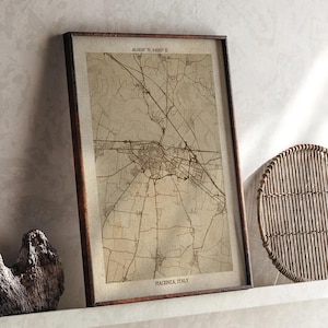 May include: A framed vintage map of Piacenza, Italy, in a brown wooden frame. The map shows detailed road and building layouts, with the city's name and coordinates at the bottom and top. The map is displayed on a white shelf.